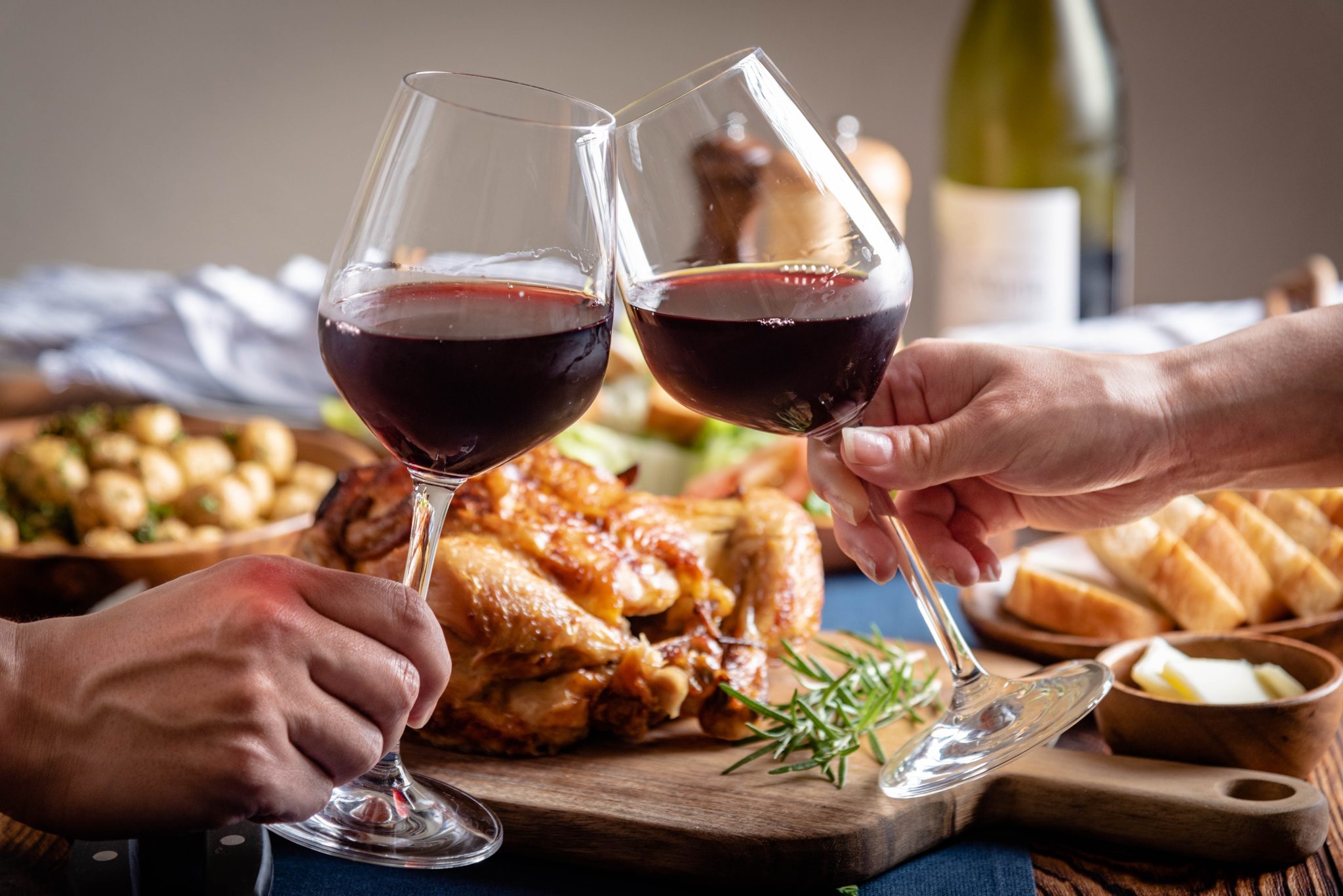 A shot of two red wine glasses toasting with roasted chicken behind