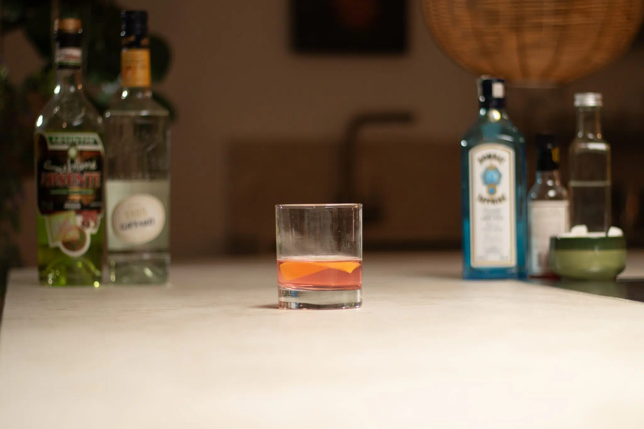 Gin, Triple Sec, sugar syrup, sugar cubes in a bowl, Peychaud's bitters, and Absinthe laid out on a white bar table