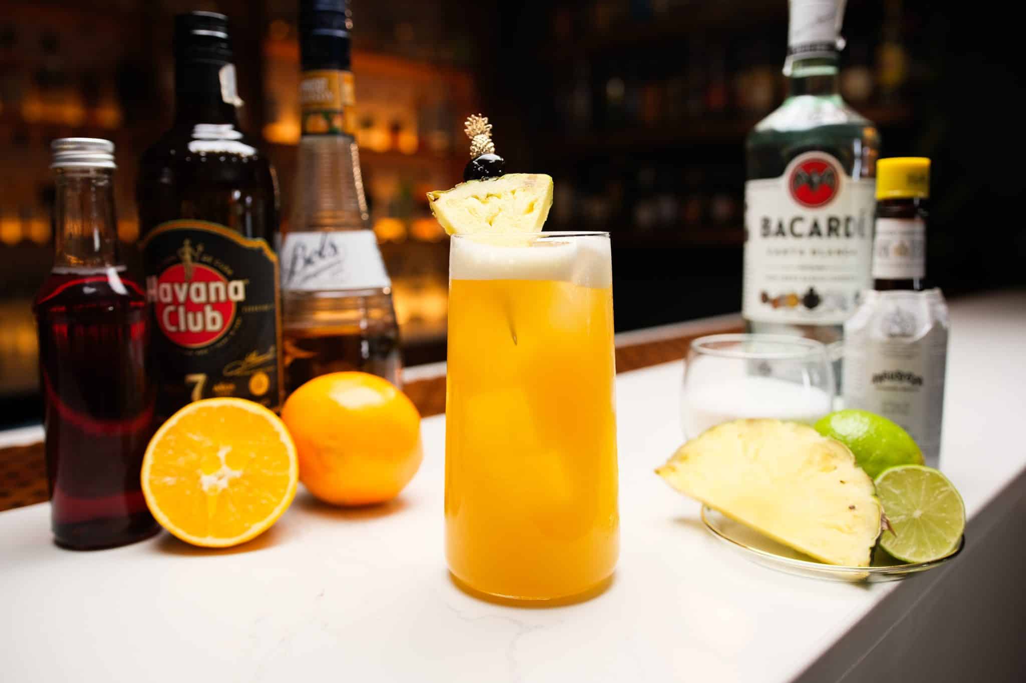 White rum, dark rum, apricot brandy, grenadine, angostura bitter, oranges, pineapple, limes and superfine sugar laid out on a white bar table.