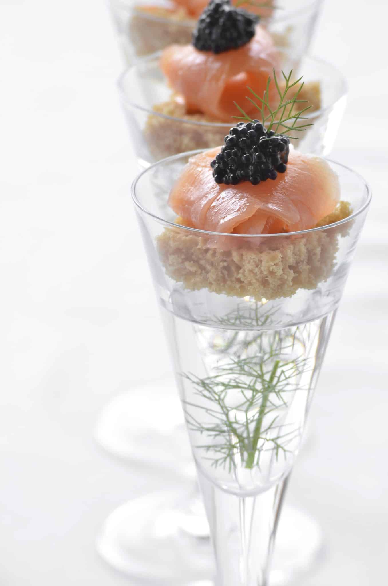 Three glasses with aquavit and sprig of dill, layered with brown bread and topped with salmon and black caviar, on a bright background.