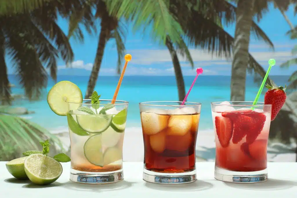 A side shot of three cocktails on a white table with a carribean beach as background.