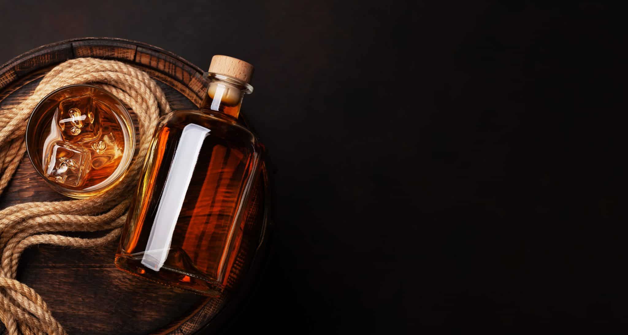 A glass of brandy with ice next to a bottle, on a wooden barrel with a rope, against a dark background.