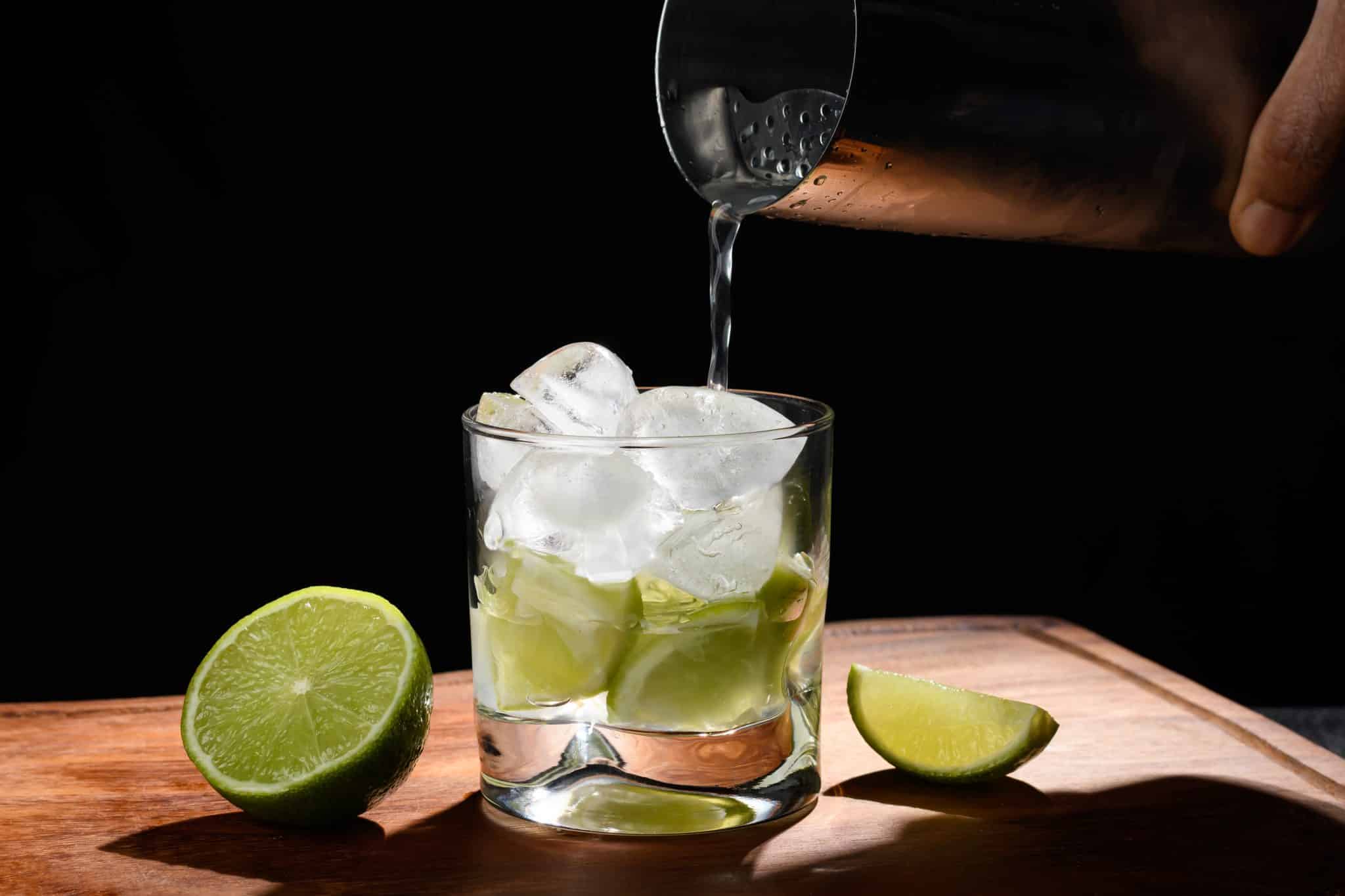 Pouring cachaça into a glass with ice and lime wedges on a wooden surface, with a dark background.
