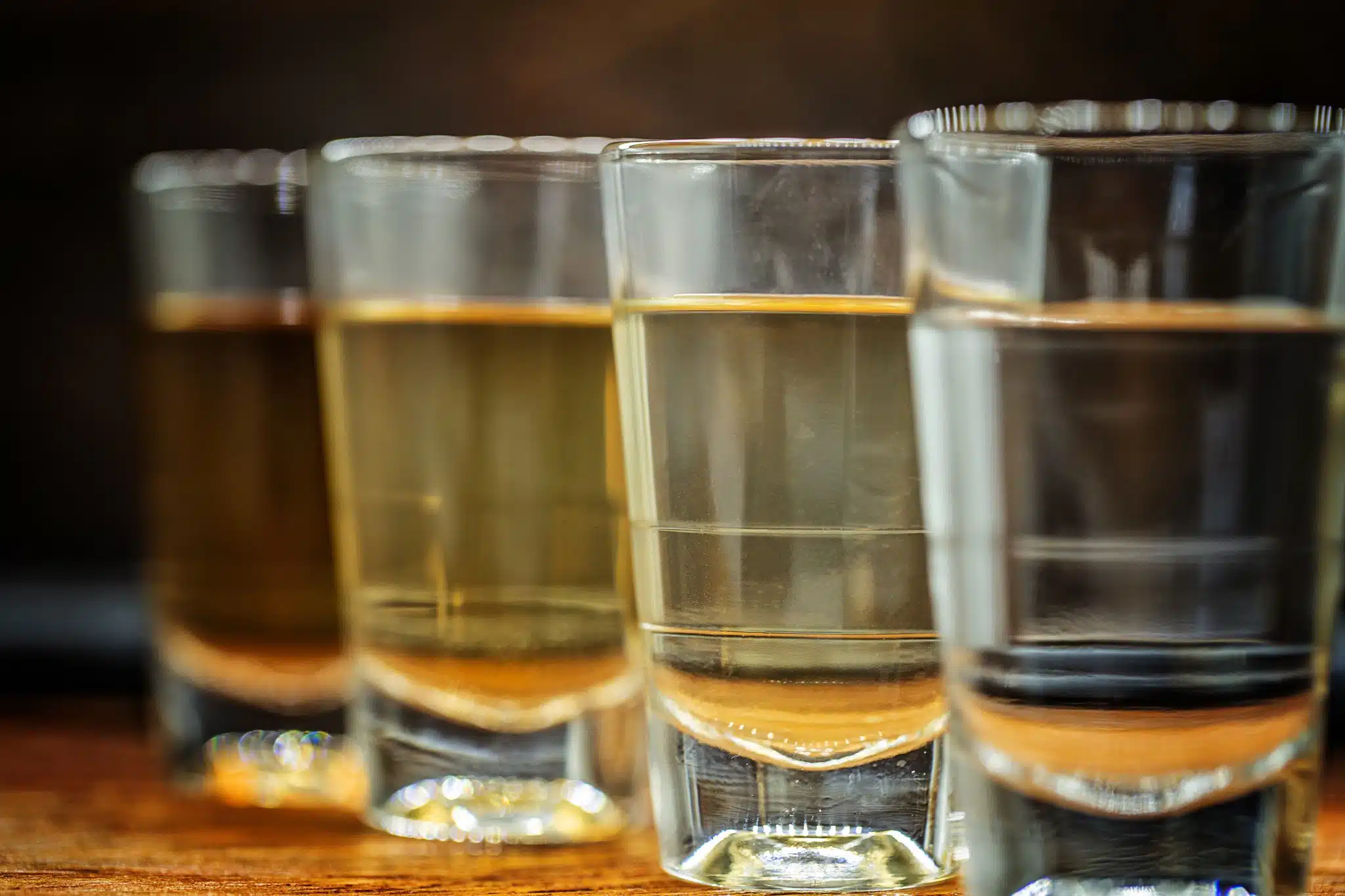 Several small glasses filled with golden liquid, lined up on a wooden surface with a dark background, likely containing cachaça, a Brazilian spirit.