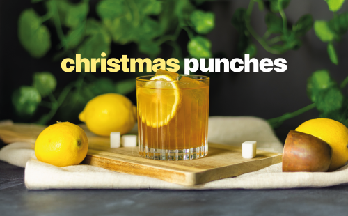 A Christmas cocktail in an old fashioned glass on a wooden board on a grey surface surrounded by three lemons, three sugar cubes and a white cloth