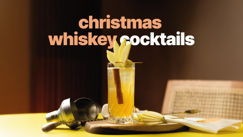 A side shot of a whiskey cocktail in a highball glass on a wooden board surrounded by a cocktail shaker, a bar spoon, apple slices and a white cloth