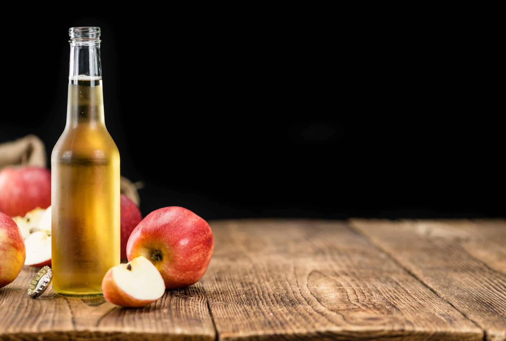 The image shows a clear bottle of golden liquid on a wooden table with red apples around it, suggesting apple cider, with a dark background.