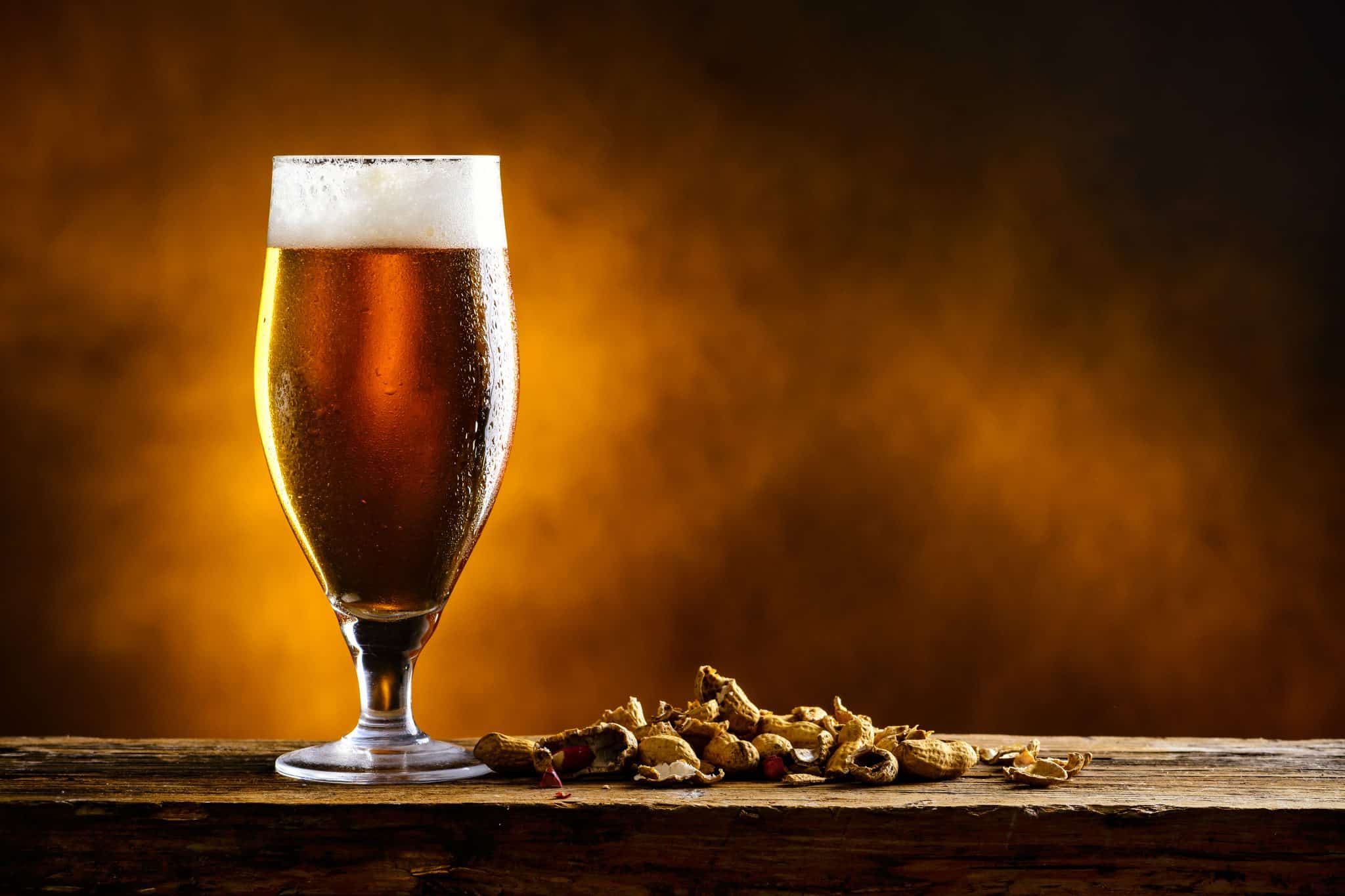 The photo shows a tall glass of beer with foam on top, on a wooden surface with peanuts next to it, and a warm, dark background.