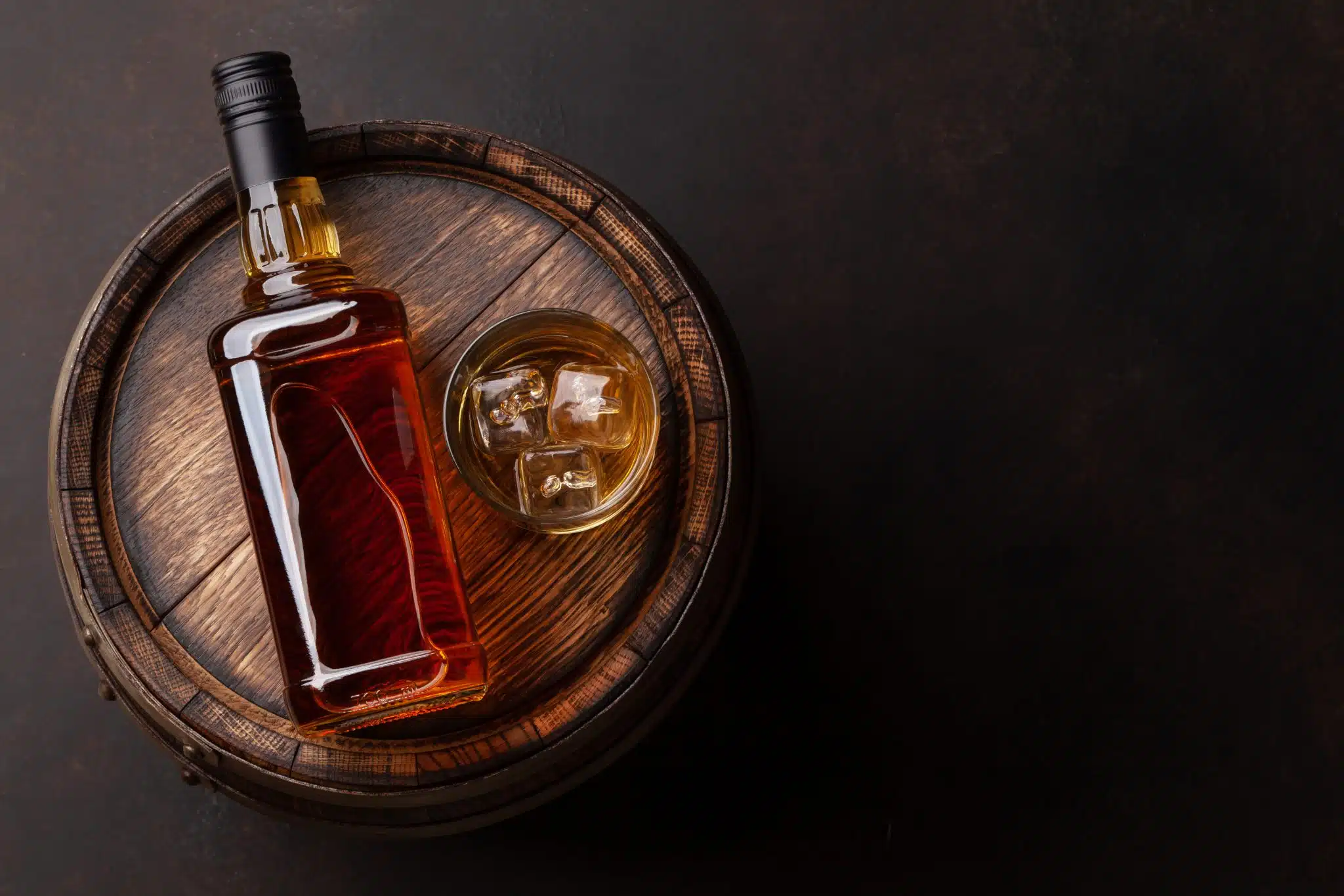 A bottle and a glass with dark liquor, shot from above, on a barrel with a black blackground.