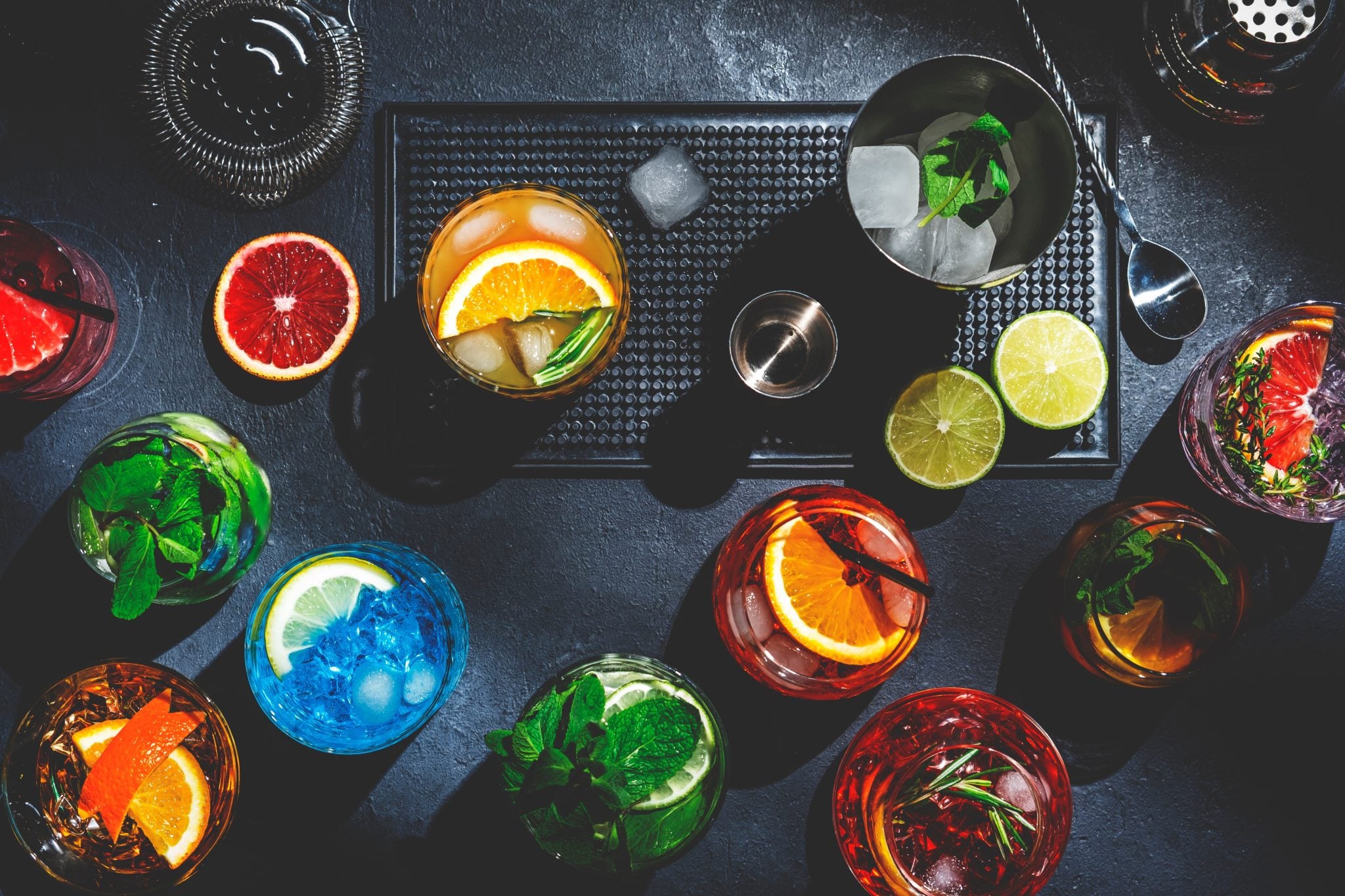 A shot from above of different types and colors of mocktails from on a dark grey surface with some ice cubes and citrus fruits around