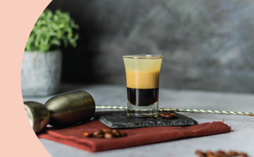 A side shot of a layered shot cocktail in a shot glass on a black stone placed on a light grey surface surrounded by a red cloth, a jigger, a bar spoon, and coffee beans