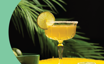 A side shot of a rimmed coupe glass with a yellow cocktail on a yellow surface with a green bowl on the site with two limes inside, in front of a black background