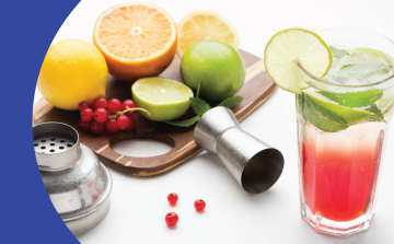 A shot of a cocktail in a glass on a white surface surrounded by a jigger, a cocktail shaker lid and a wooden board with an orange, two lemons, two limes, cranberries