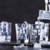 4 shot glasses being filled with ice cubes and chilled vodka on a black stone plate placed on a black table in front of a black background.