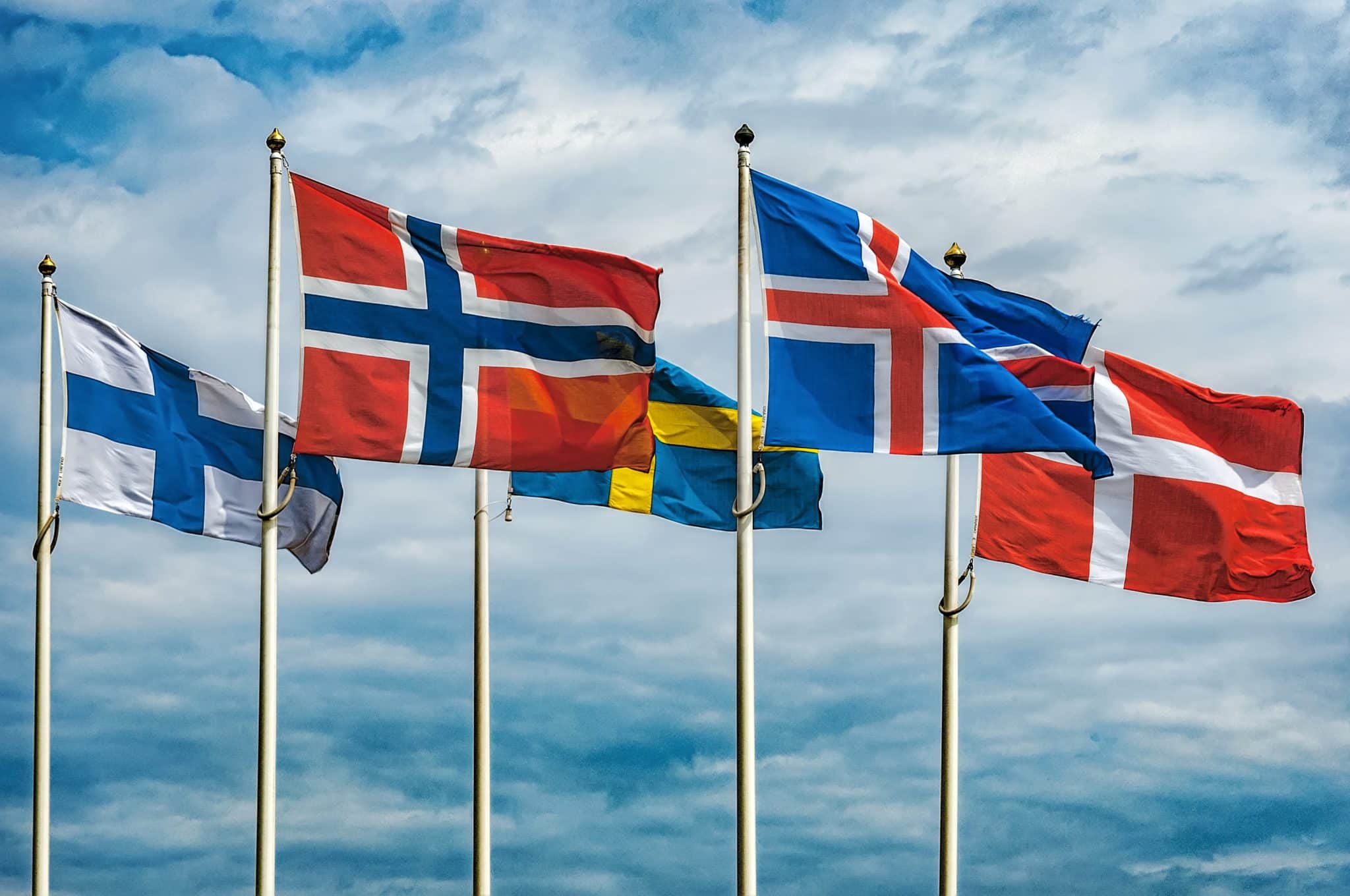 Five Nordic flags waving against a cloudy sky, representing Finland, Norway, Sweden, Iceland, and Denmark from left to right.