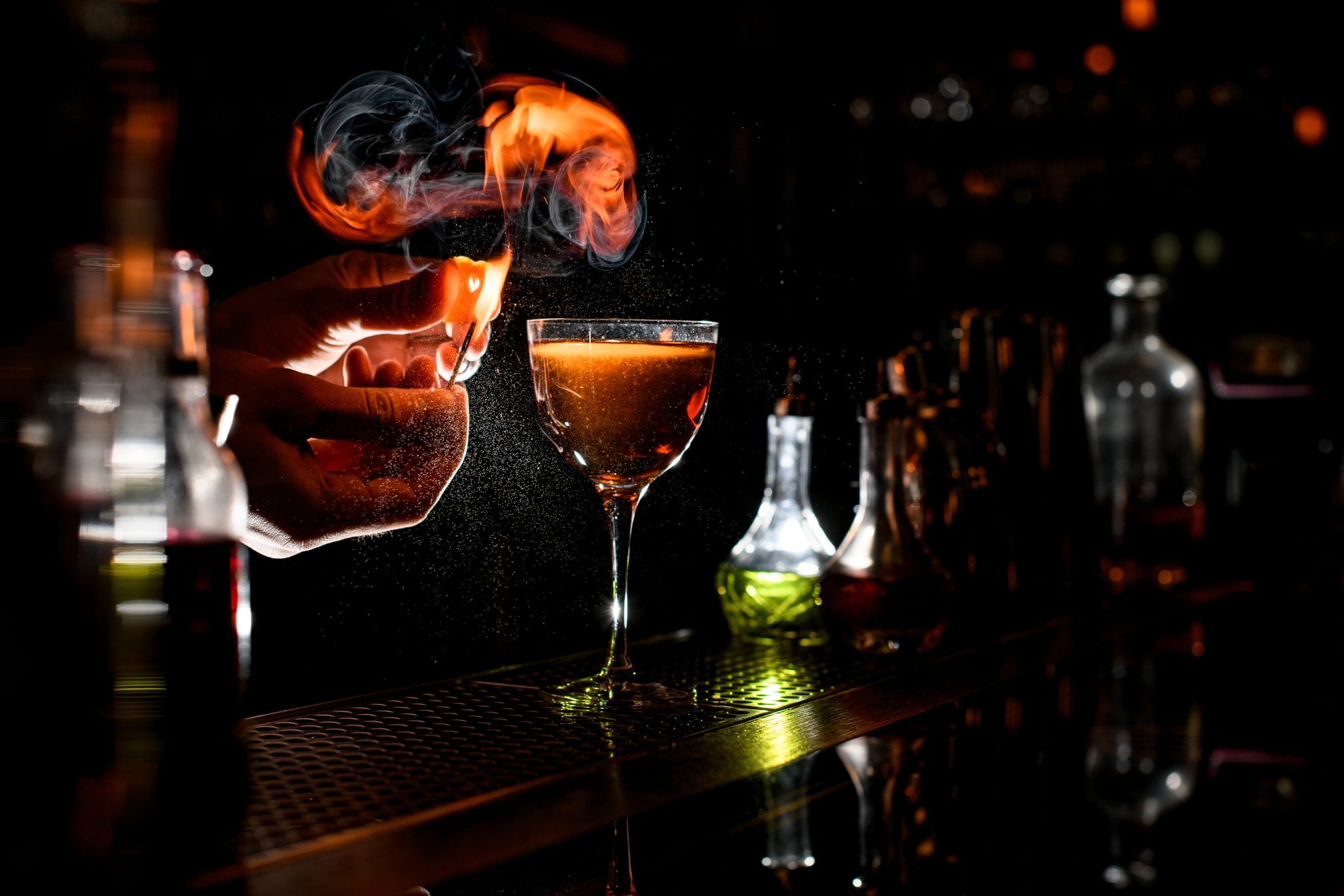 Shot of a bartender expressing the oils of a citrus twist and holding a flame close to it and to a cocktail on a bar table