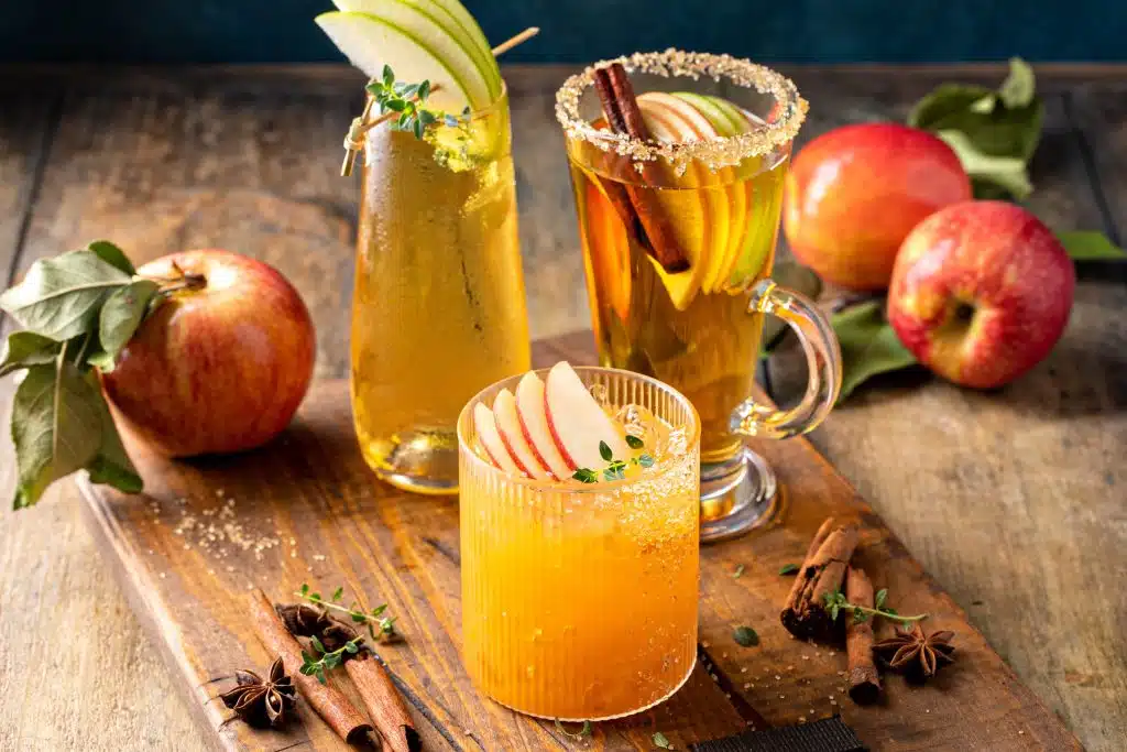A shot of three fall cocktails on a wooden table with cinnamon barks and apples around.