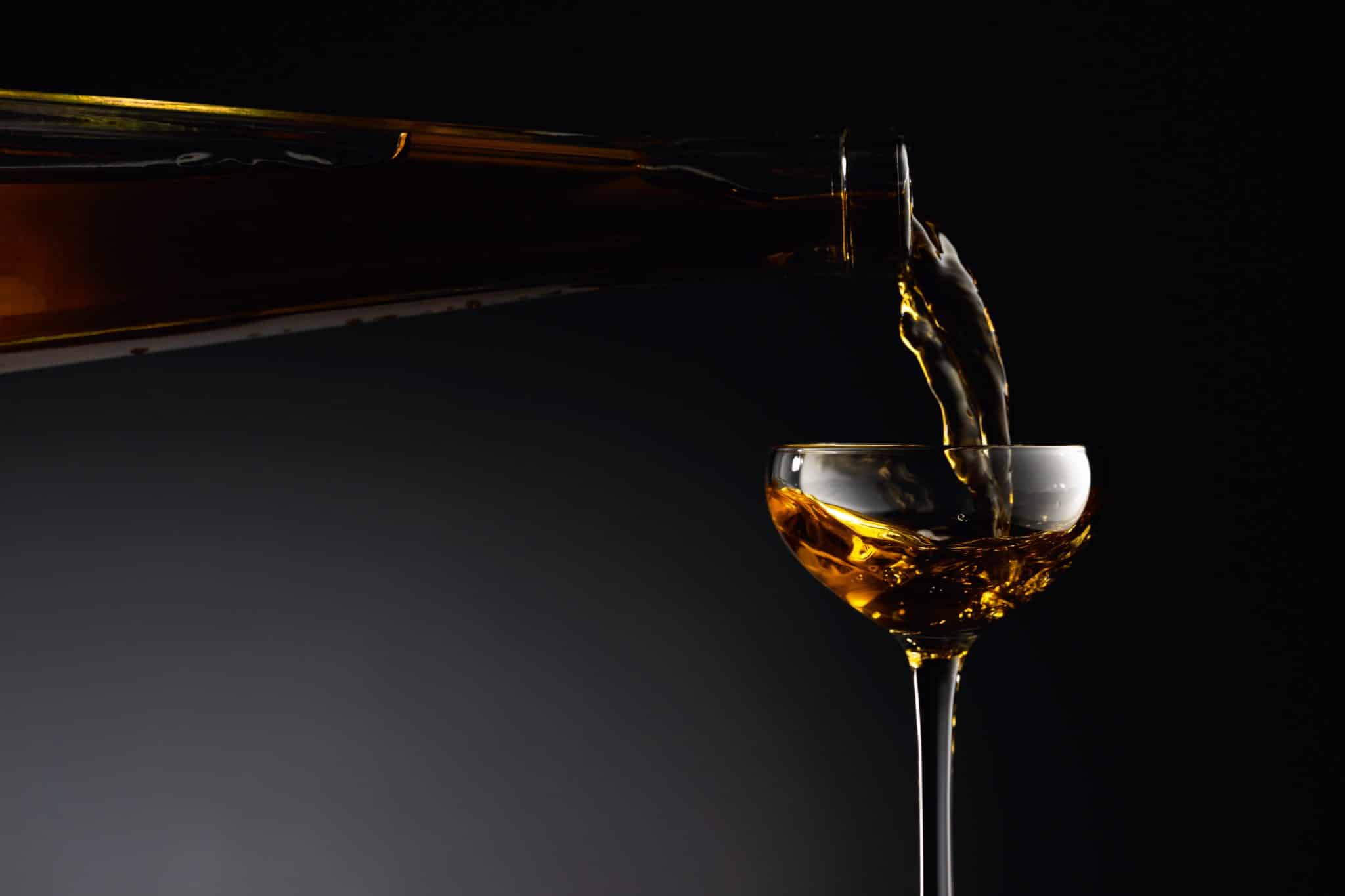 The image shows a bottle tilted and pouring a fortified wine into a wine glass against a dark background.