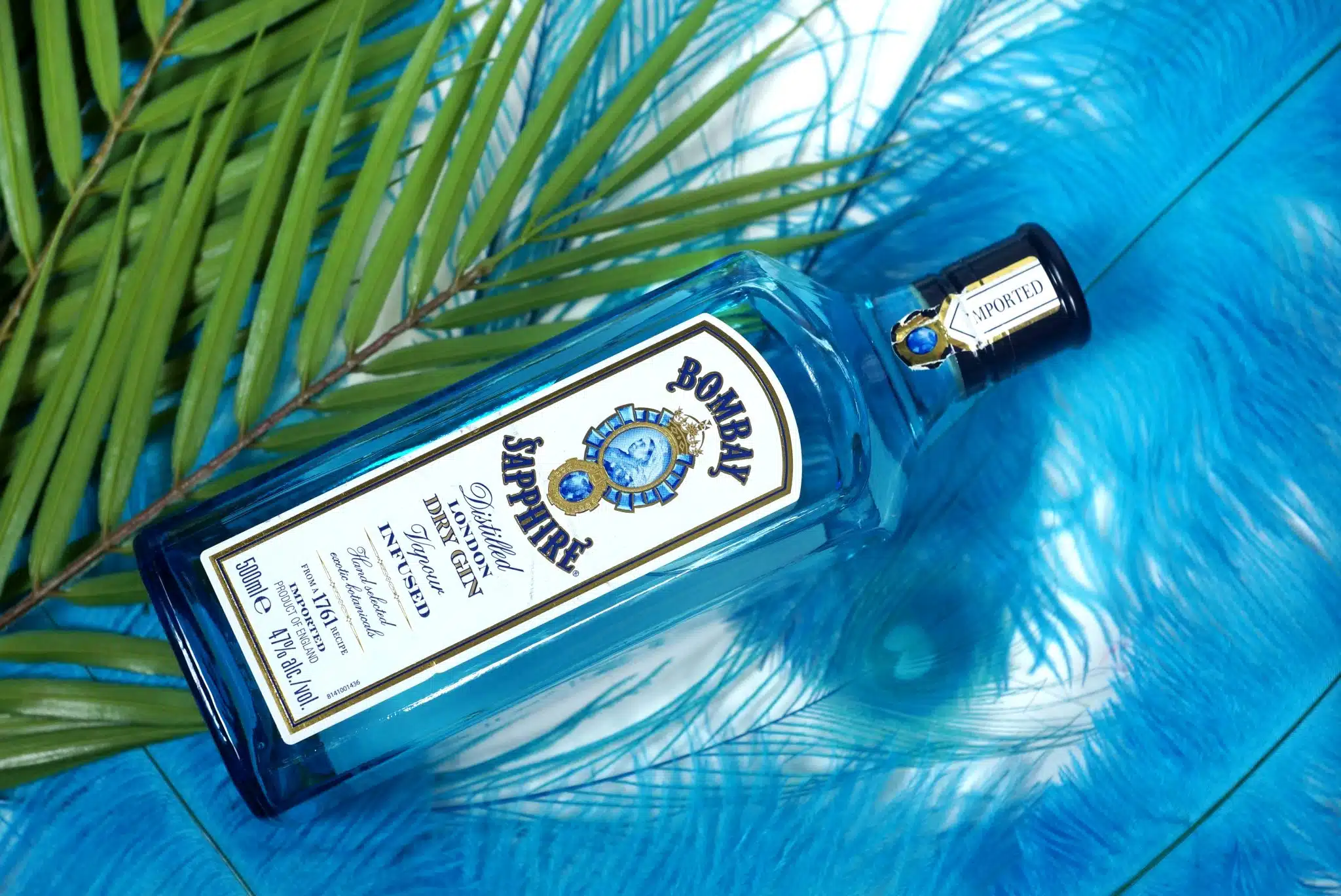 A Bombay Sapphire gin lying on a blue surface with green palm fronds.