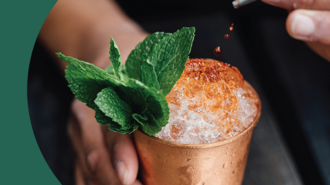 A shot of a cocktail in a julep cup garnished with mint and balanced with some drops