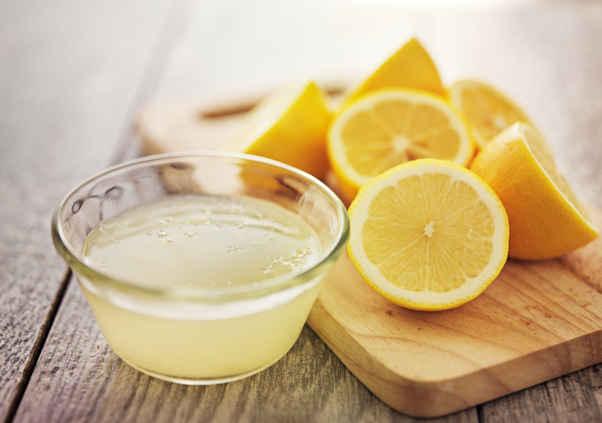 A shot of lemon juice in a bowl with five half lemons on a wooden board on a side