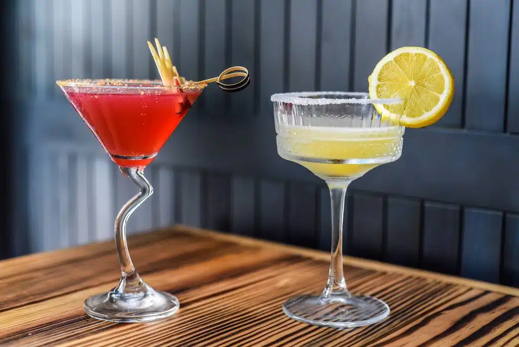 A Cosmopolitan and a Margarita cocktails on a wooden table on a dark blue blackground.