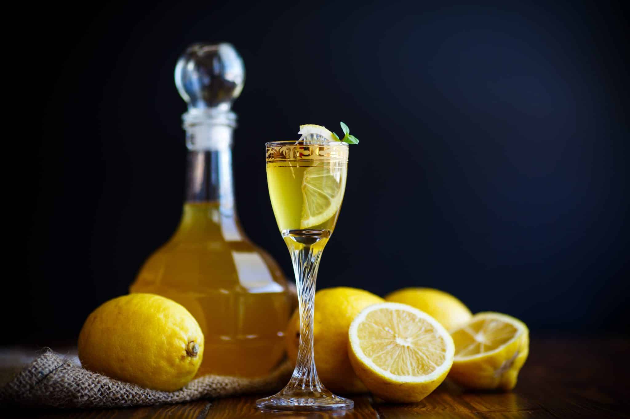 A glass of yellow liqueur garnished with a lemon slice and mint, with whole lemons and a bottle in the background on a wooden surface.