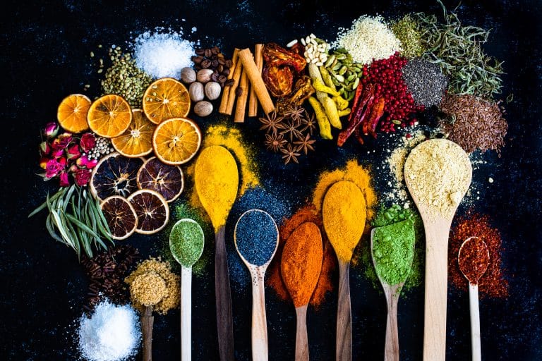 An array of colorful spices and herbs in wooden spoons on a dark background, with dried citrus and flowers, conveying a rich tapestry of flavors and scents.