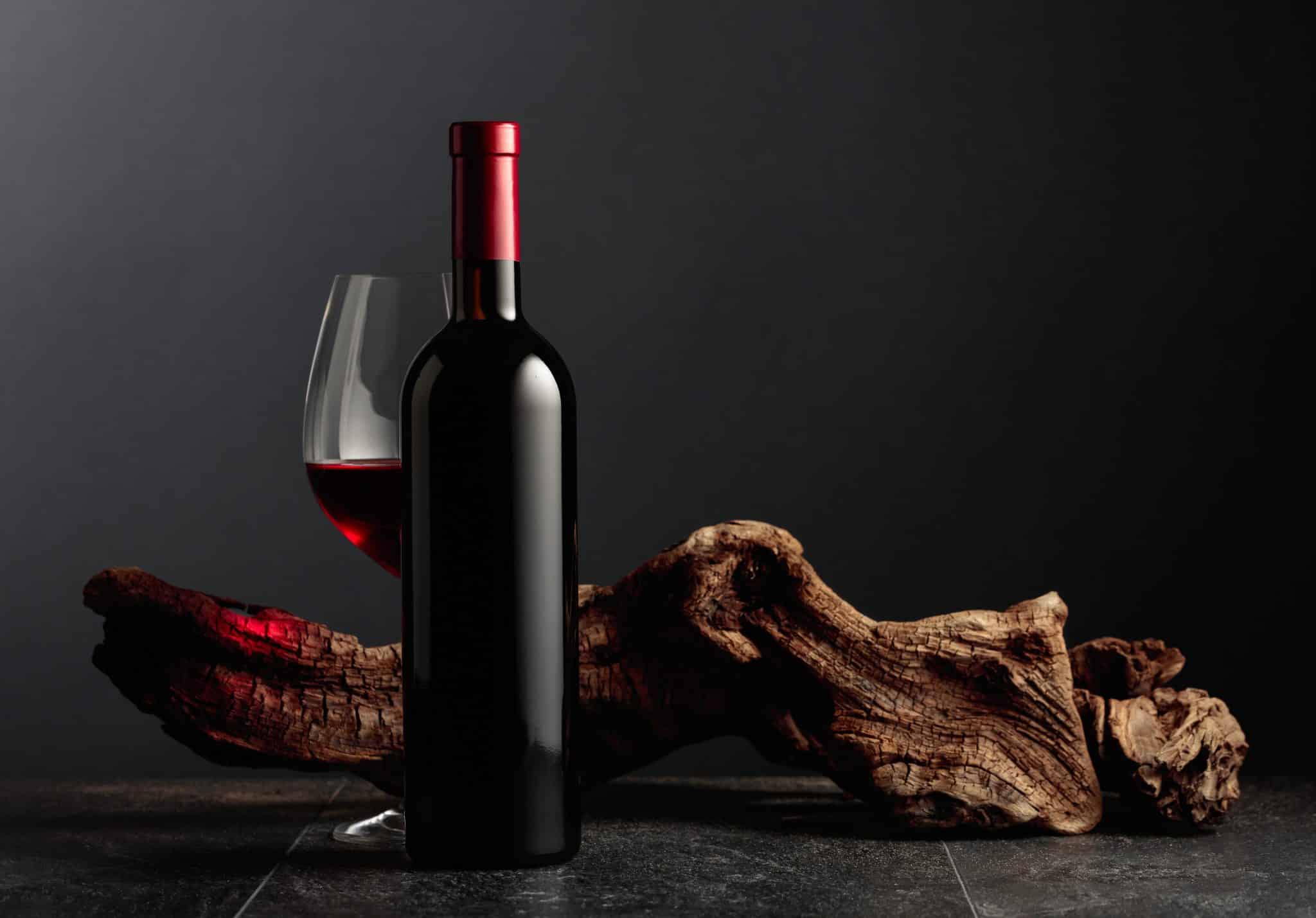 The picture shows a bottle of red wine next to a full glass, with a piece of decorative wood in the background, all set against a dark backdrop.