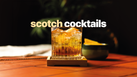 A shot of a scotch cocktail in an old fashioned glass on a wooden coaster placed on a wooden surface surrounded by a ber spoon, a white cloth, and a green bowl with orange slices