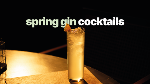 A side shot of a cocktail in a colling glass garnished with an orange peel placed on a round surface, in front of a black background