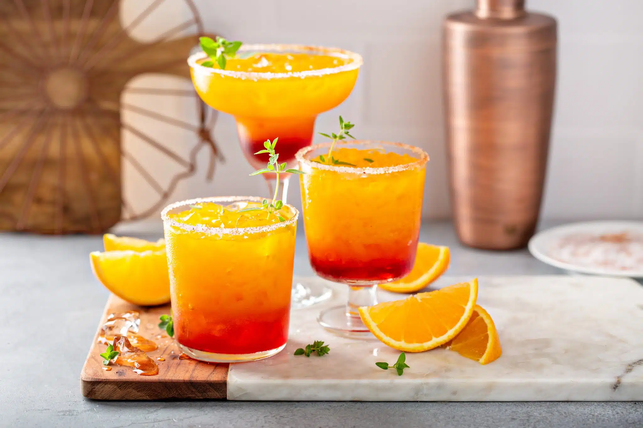 A shot of three tequila sunrise cocktails in three different glasses on a wooden-marmol tray surrounded by orange pieces and a shaker on the background.